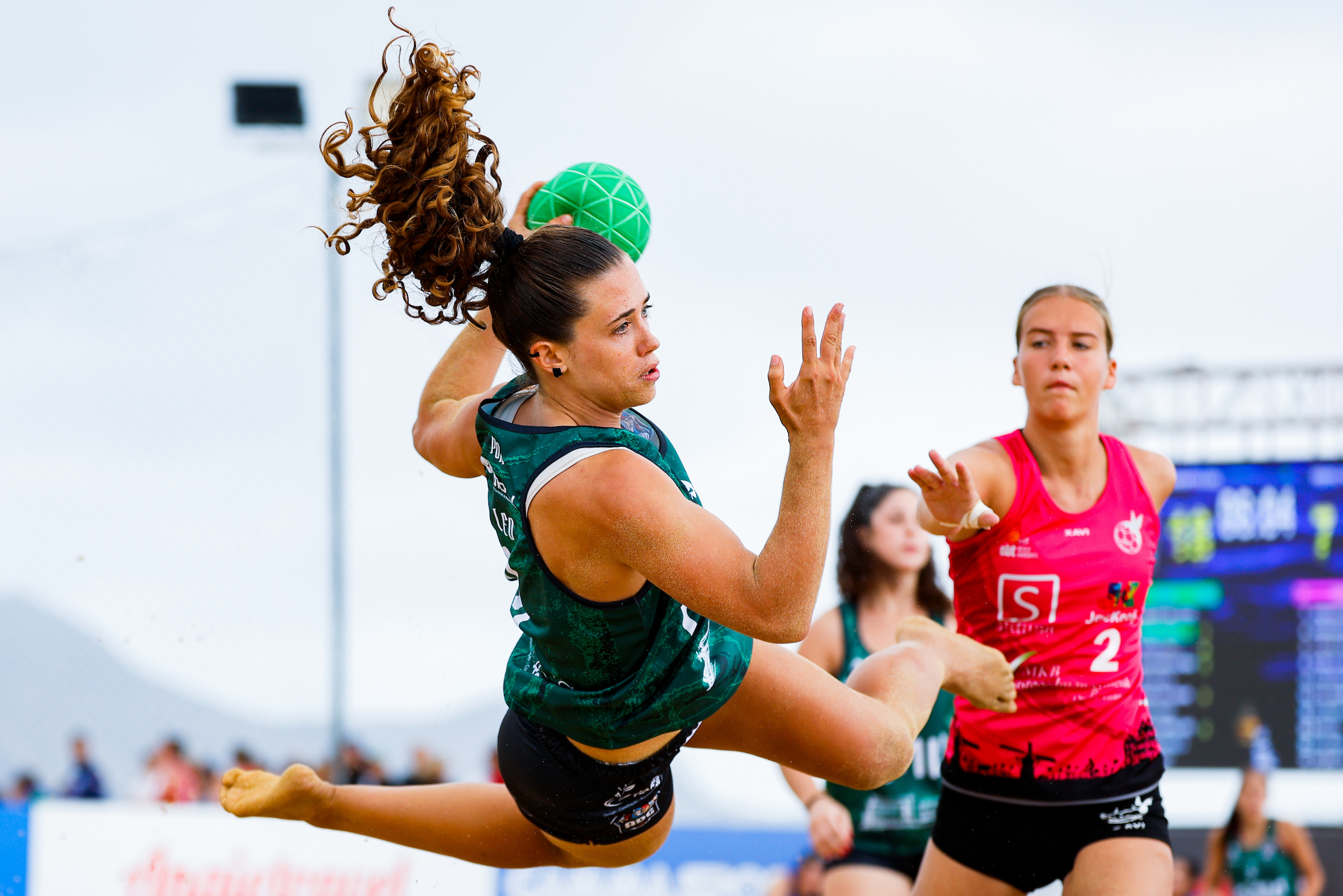An athlete in a green uniform leaps mid-air to catch a green ball during a fast-paced outdoor sports match, while an opponent in a red jersey watches closely.