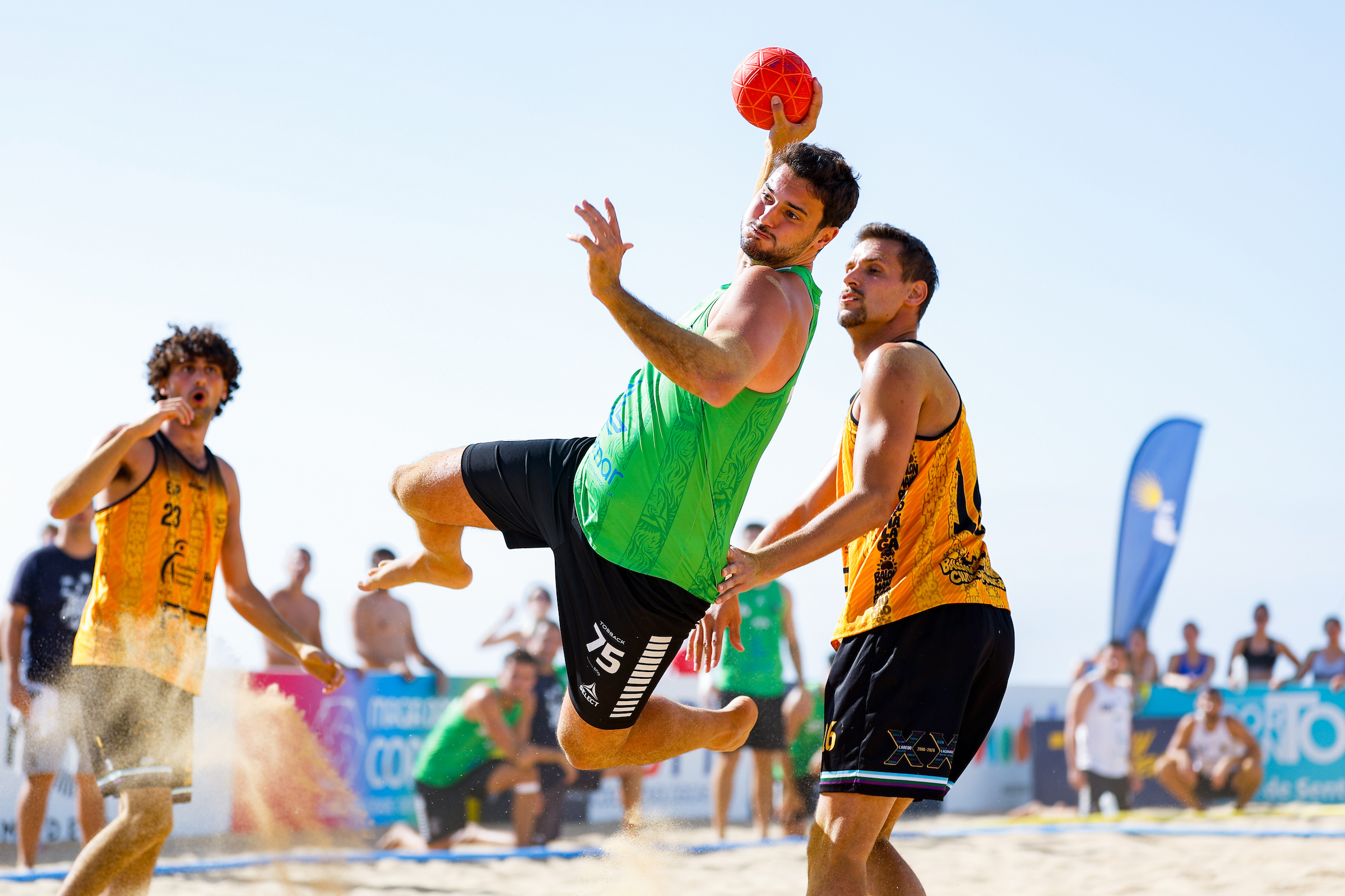 An athlete in a green jersey leaps to catch a red ball while being defended by an opponent on a sandy beach court.